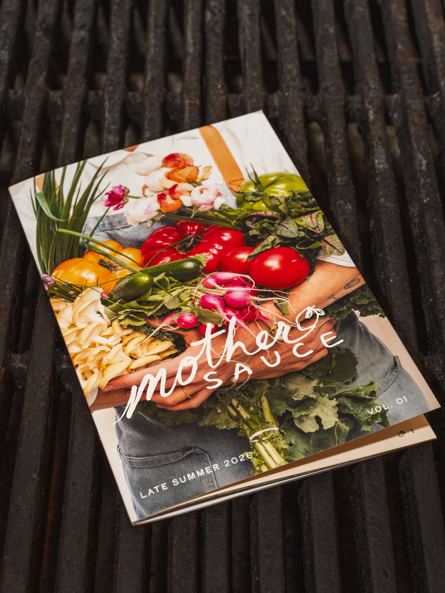 A printed magazine or cookbook titled "mother's sauce" lies on a dark wooden slatted surface. The cover features a colorful photo of fresh vegetables and pasta, with the title in large white script.